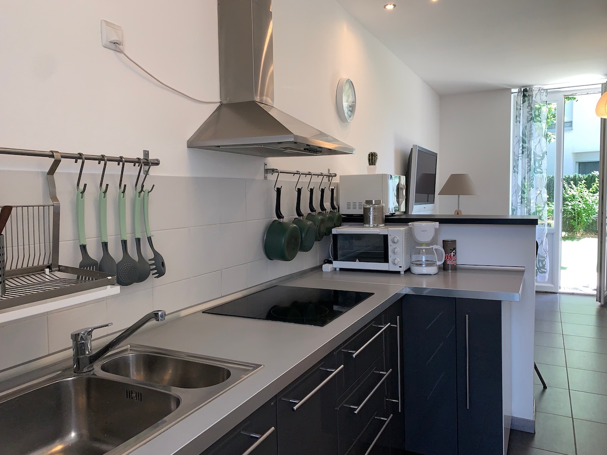 The kitchen area showcases modern appliances, including a microwave and a stovetop. A stainless steel sink is positioned alongside a functional counter space. Cooking utensils and cookware are neatly arranged on a wall rack. Natural light filters in through a nearby window, enhancing the space.