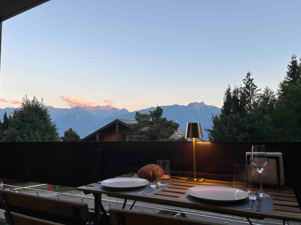 A dining table is set on a terrace, featuring two plates, a loaf of bread, and glasses. A lamp provides soft illumination, while the backdrop showcases distant Swiss Alps under a pastel sky.