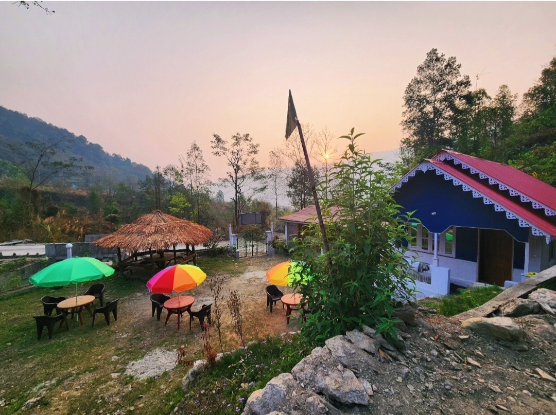 An outdoor seating area features multiple round tables surrounded by colorful umbrellas, set against a backdrop of lush greenery and mountains. A thatched-roof structure is visible in the distance, with the warm hues of sunset illuminating the scene.