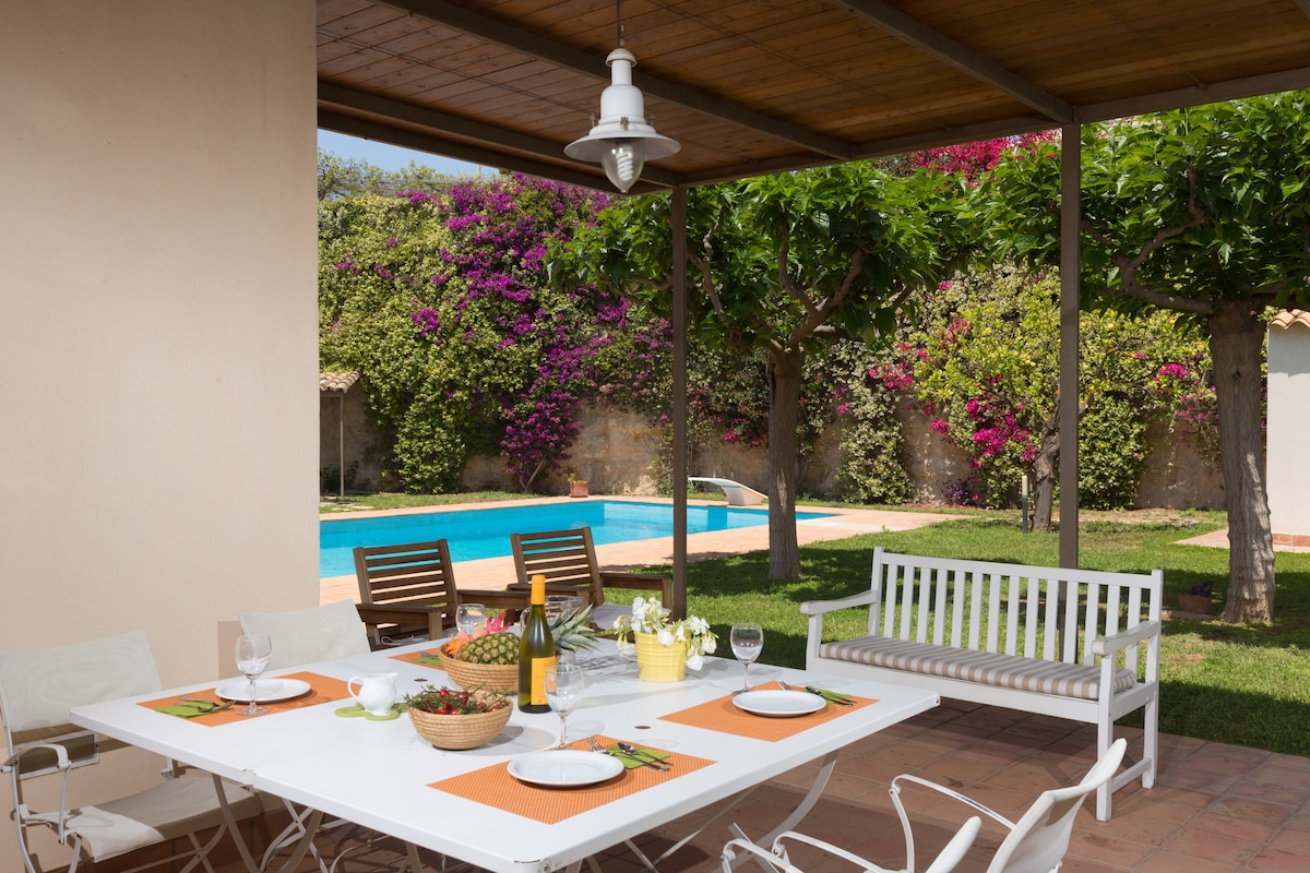 An outdoor dining area is presented under a wooden pergola. A large table is set with dishes and drinks. Nearby, a pool glimmers in the background, framed by manicured grass and flowering shrubs. Seating includes two lounge chairs and a white bench.