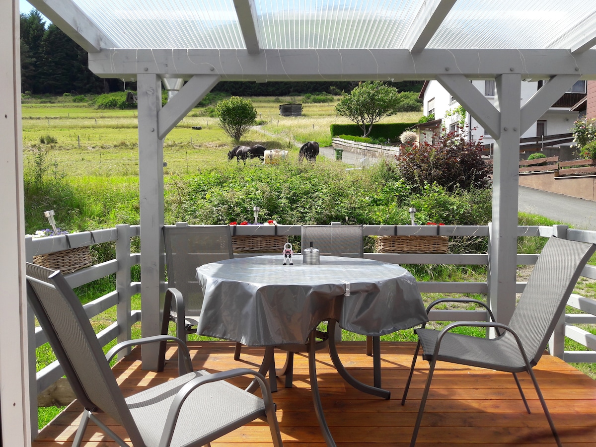 A covered outdoor terrace features a round table with a gray tablecloth and four chairs, situated in a peaceful setting. Lush greenery and open fields are visible beyond the railing, with grazing animals in the distance, providing a serene and picturesque view.