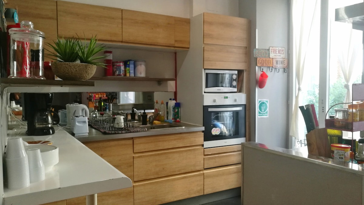 A modern kitchen area is seen with wooden cabinetry and a white countertop. Kitchen appliances, including a coffee maker and oven, are visible. A variety of colorful items are placed on the shelves, and natural light enters through large windows, enhancing the space.