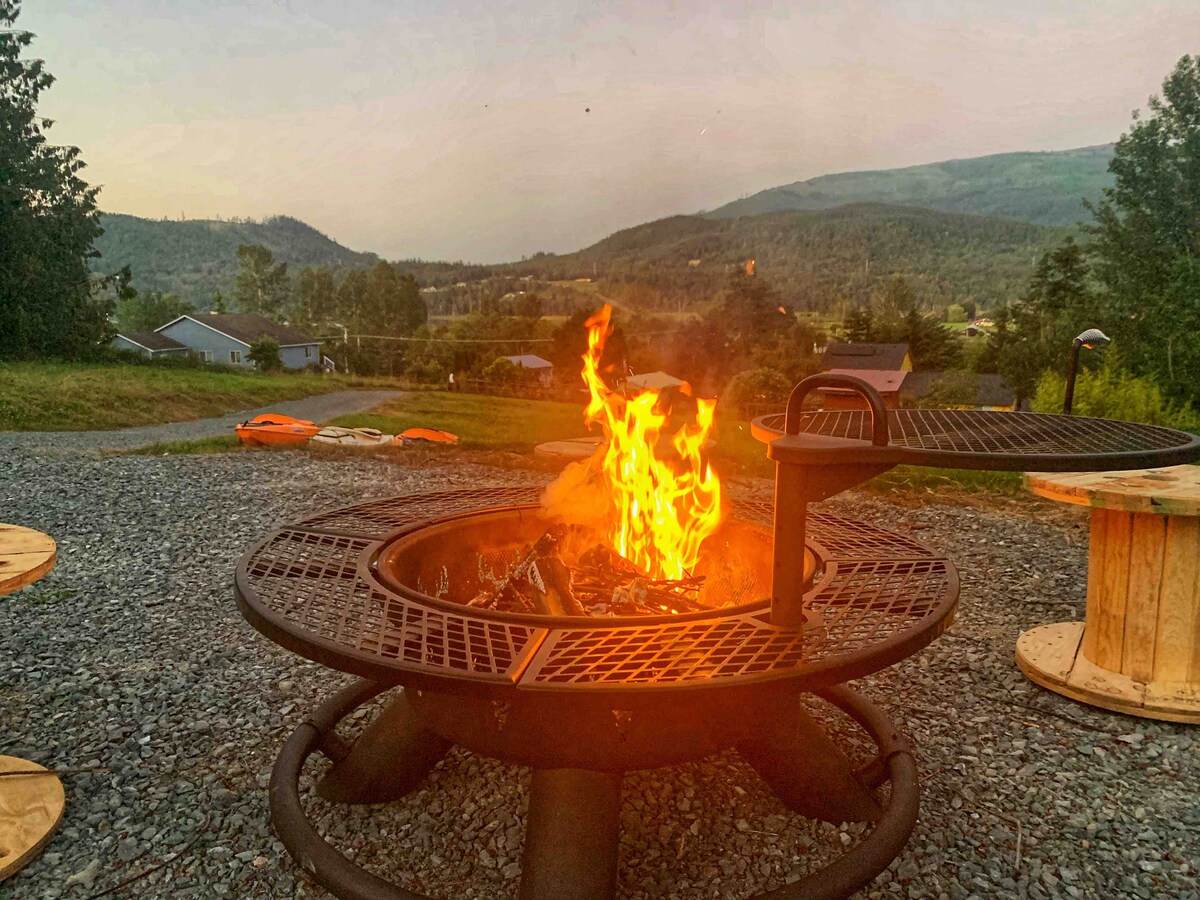 A fire pit is centrally placed on a gravel area, surrounded by a few wooden seating arrangements. Flames are captured dancing upward against a backdrop of rolling hills and mountains during twilight.