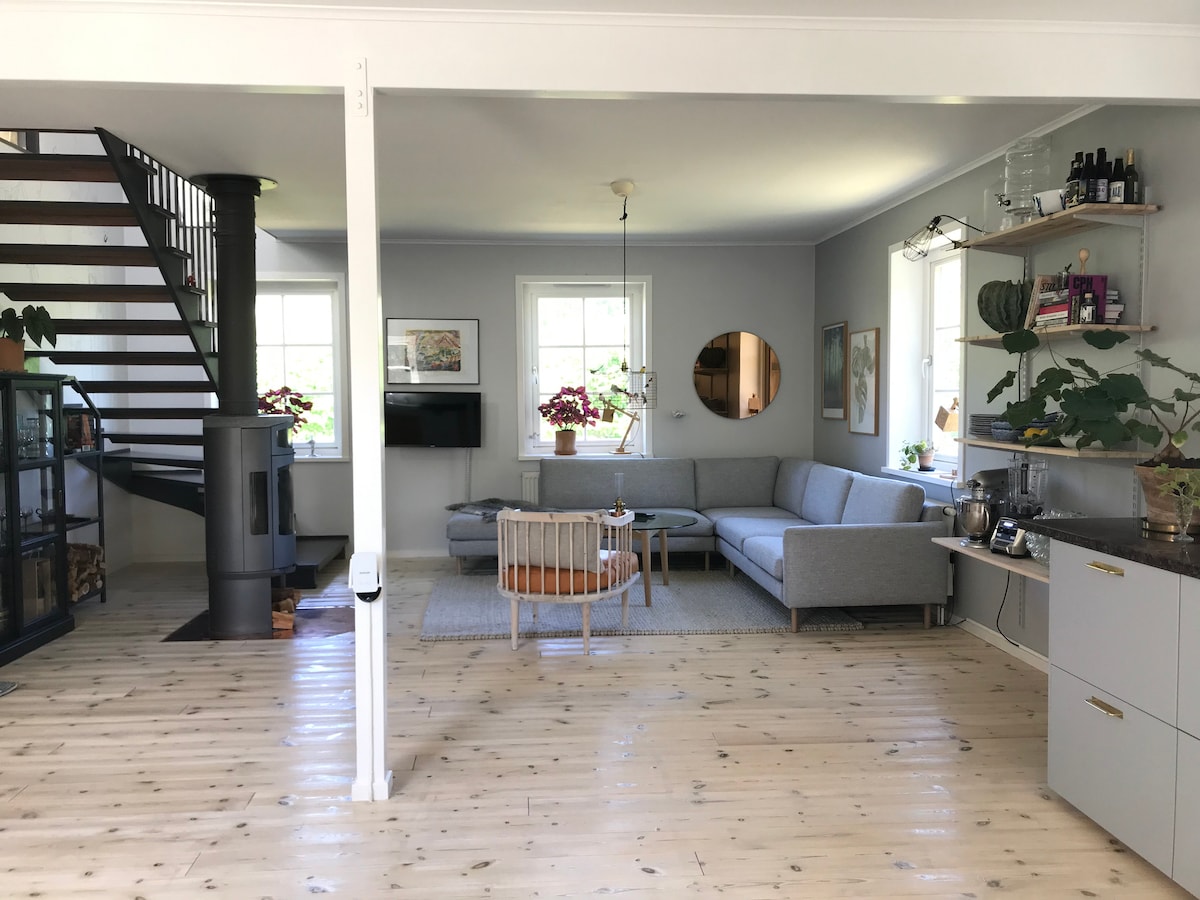 A spacious living area is presented, featuring a light grey sectional sofa and a round coffee table. Natural light is allowed by multiple windows, and a staircase is visible in the background. Decorative plants and art add a personal touch to the neutral decor.