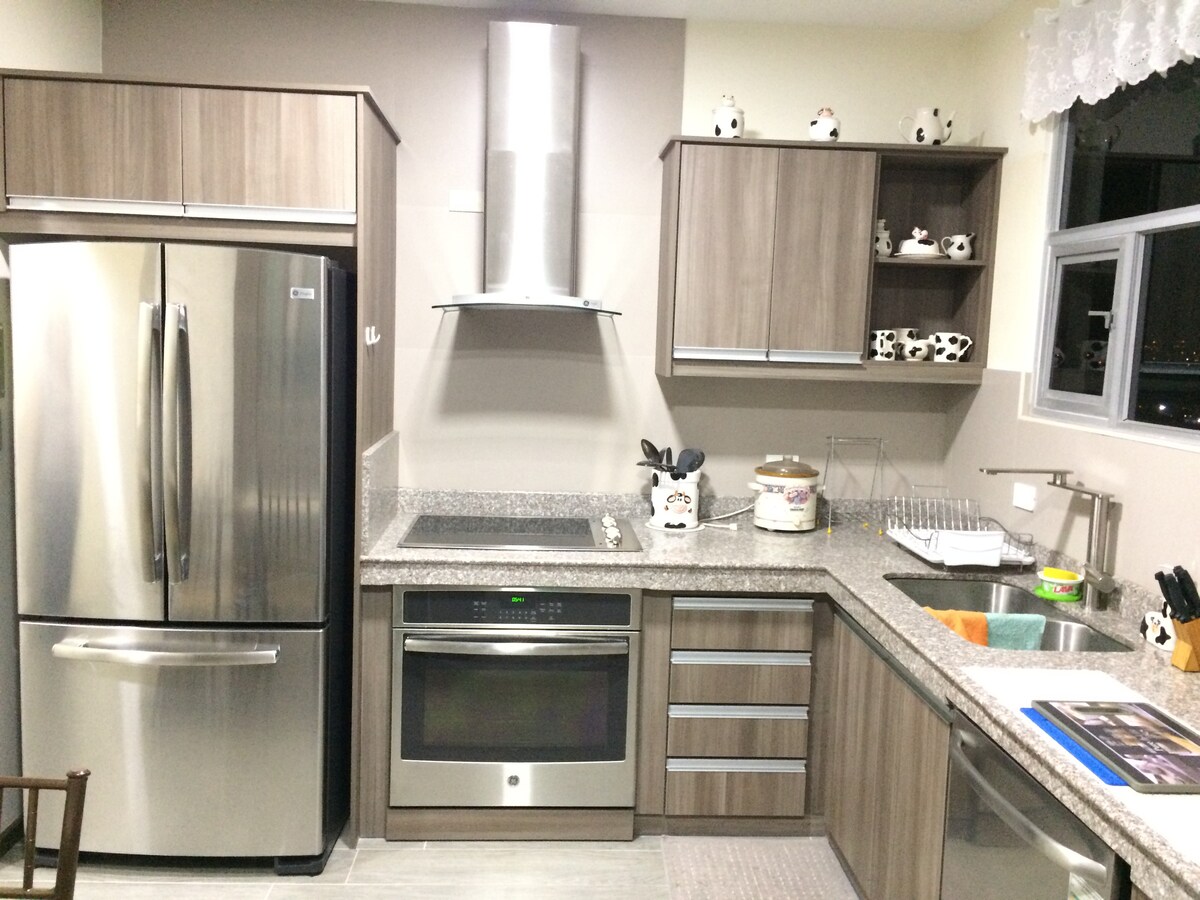 A modern kitchen features sleek wooden cabinetry and a stainless-steel refrigerator. A granite countertop is complemented by an oven and a sink. Cooking utensils and dishware are neatly arranged, showcasing a functional and well-equipped space for meal preparation.