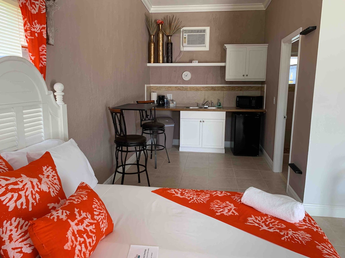 A spacious bedroom features a comfortable bed adorned with vibrant coral-patterned pillows. In the background, a kitchenette is equipped with a mini fridge and microwave, complemented by two bar stools. Warm light illuminates the room, enhancing the island-inspired decor.
