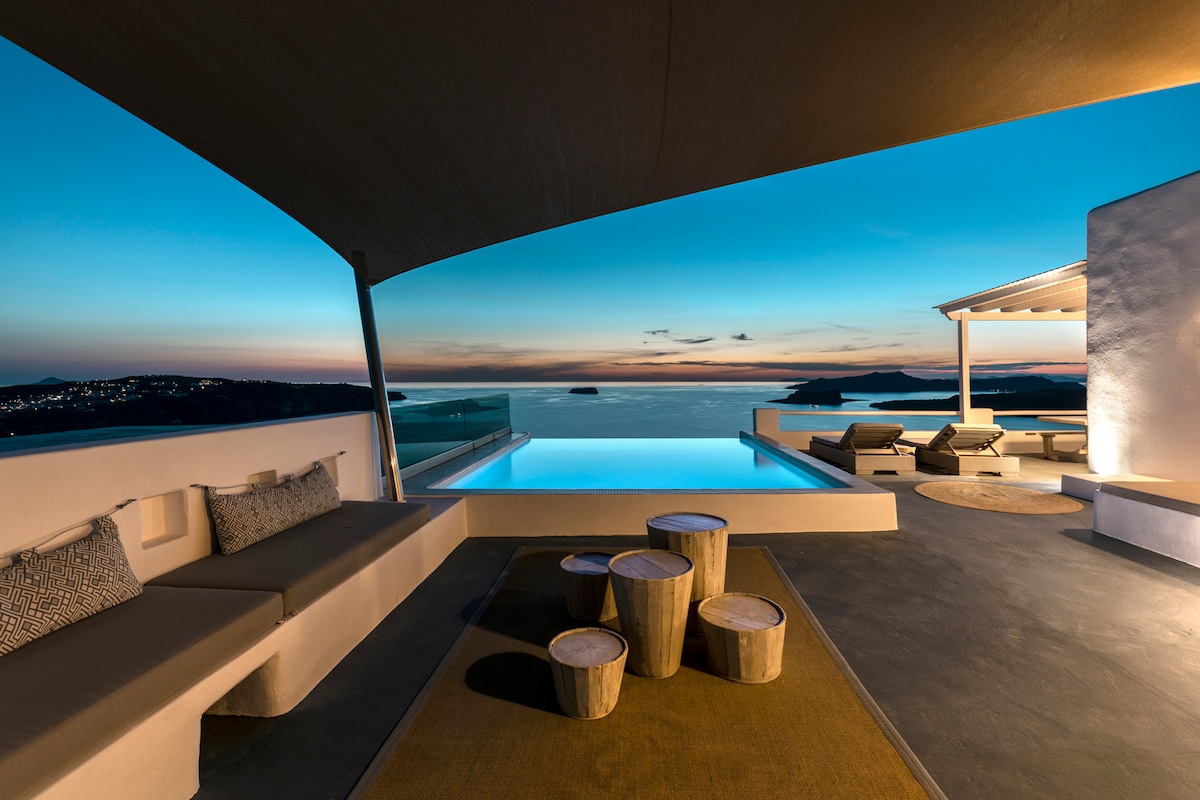 An outdoor patio features a comfortable seating area with soft cushions, accompanied by a round wooden table. A heated private pool reflects the evening sky, extending toward a picturesque view of the sea. Sunbeds and a shaded area invite relaxation during sunsets.