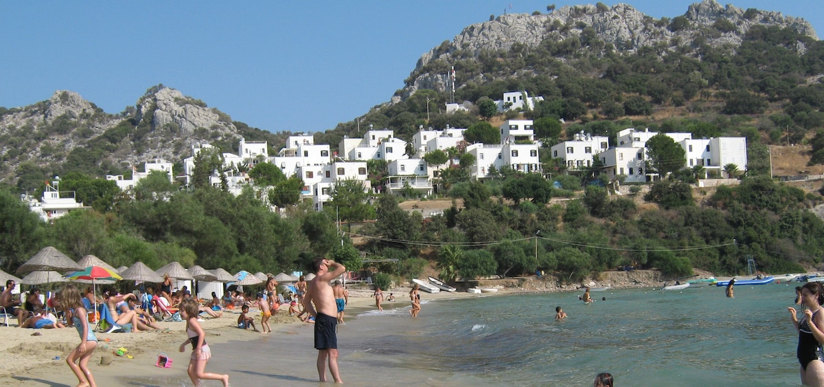 A sandy beach is lined with people enjoying the sun and water. In the background, a series of white buildings are nestled against a rocky hillside, surrounded by greenery. Umbrellas provide shaded areas on the beach, creating a lively yet relaxed atmosphere.