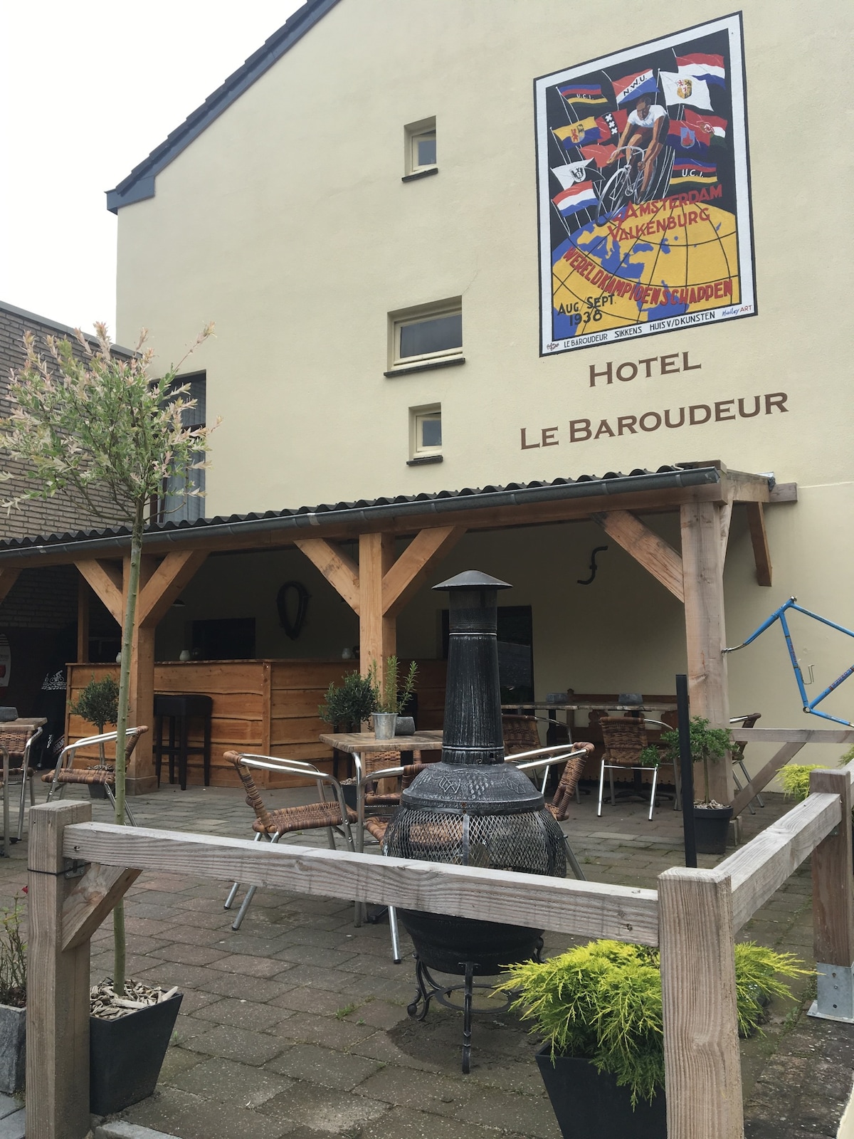 An outdoor seating area is arranged with several wooden tables and chairs, surrounded by potted plants. A large metal stove is positioned centrally, and a mural decorates the upper wall, adding character to the space. The building's architecture is complemented by wooden beams.