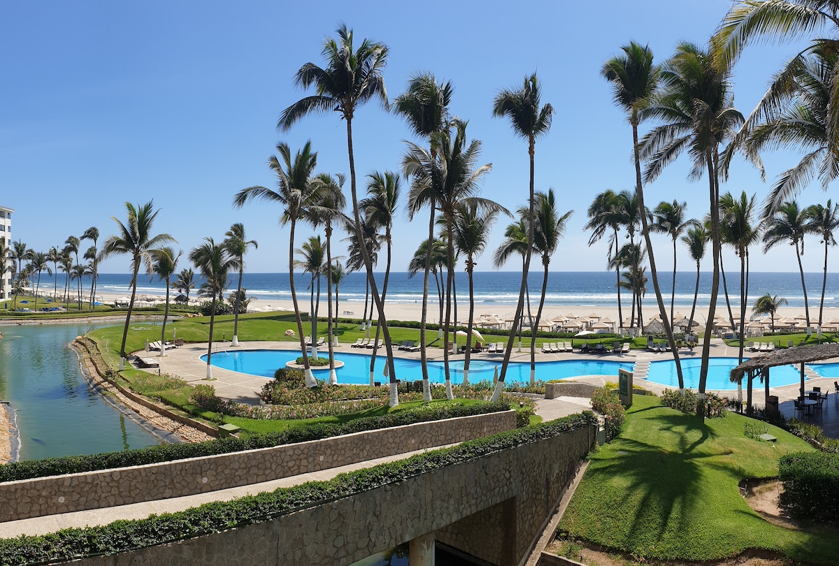A scenic view captures the manicured grounds of the resort, lined with palm trees and tropical greenery. Multiple swimming pools are visible, reflecting the sun's rays, while the ocean is seen in the background, completing the coastal landscape.