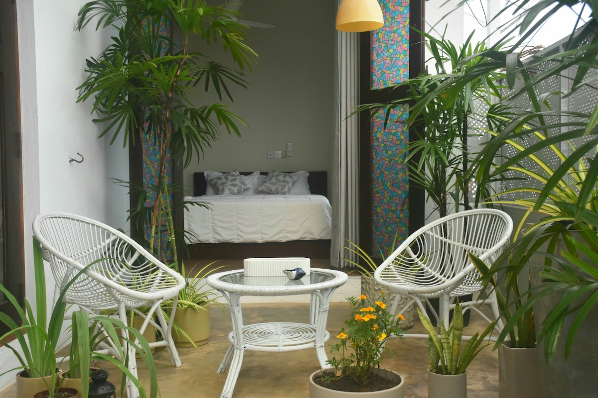 A tranquil seating area is surrounded by greenery, featuring two white woven chairs and a glass table. Potted plants, including vibrant flowers, add a touch of color. In the background, a brightly decorated wall peeks into the bedroom, enhancing the inviting atmosphere.