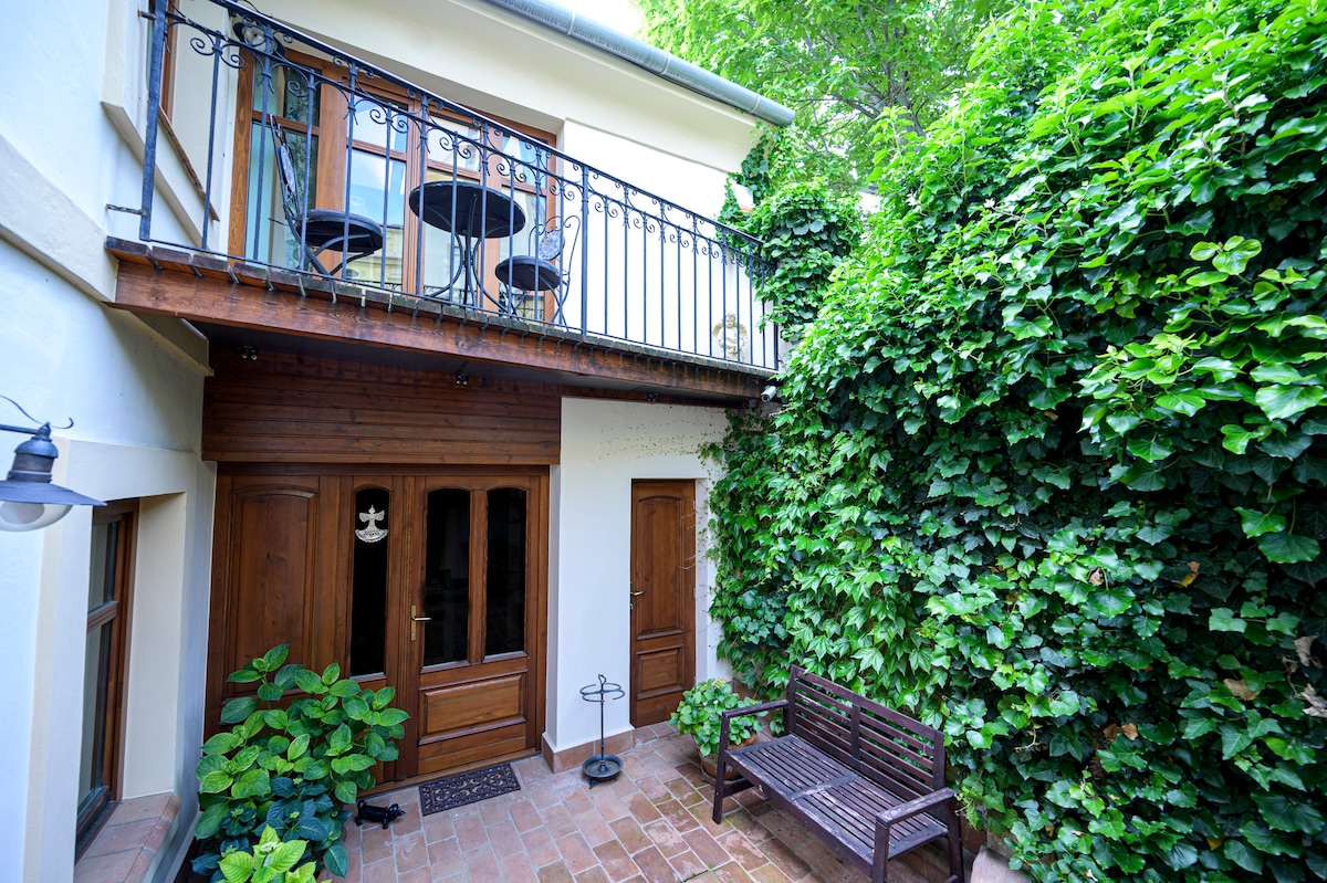 A charming courtyard is enclosed by lush green foliage, creating a serene atmosphere. Double wooden doors provide entry, while a wooden bench sits beside decorative plants. A balcony above features two small tables with chairs, inviting relaxation in a peaceful setting.