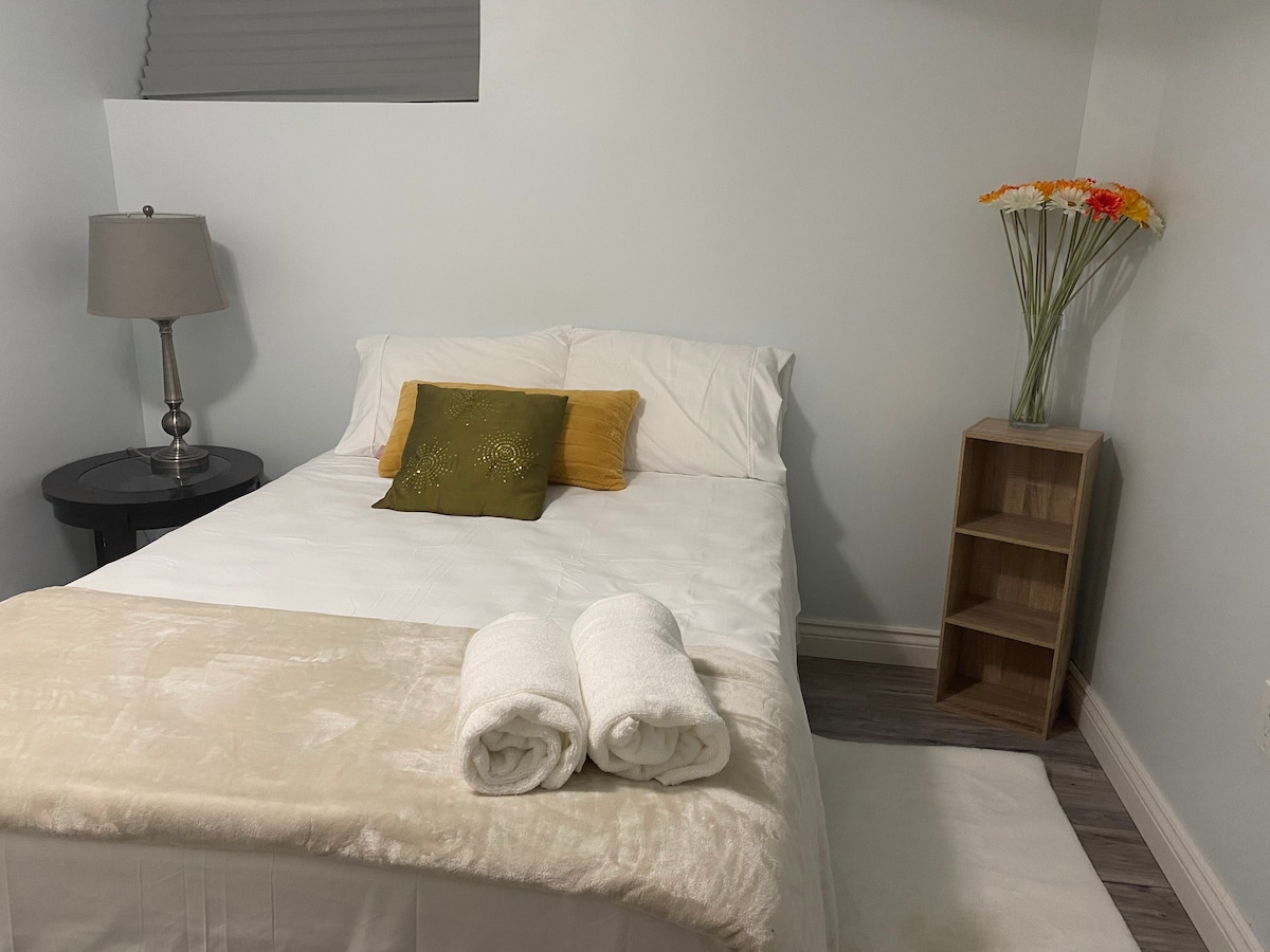 A well-appointed sleeping area features a spacious bed with neutral bedding and a decorative green pillow. Two rolled towels are placed at the foot of the bed. A bedside lamp and a wooden shelf with a vase of flowers add warmth to the room, which is softly illuminated.
