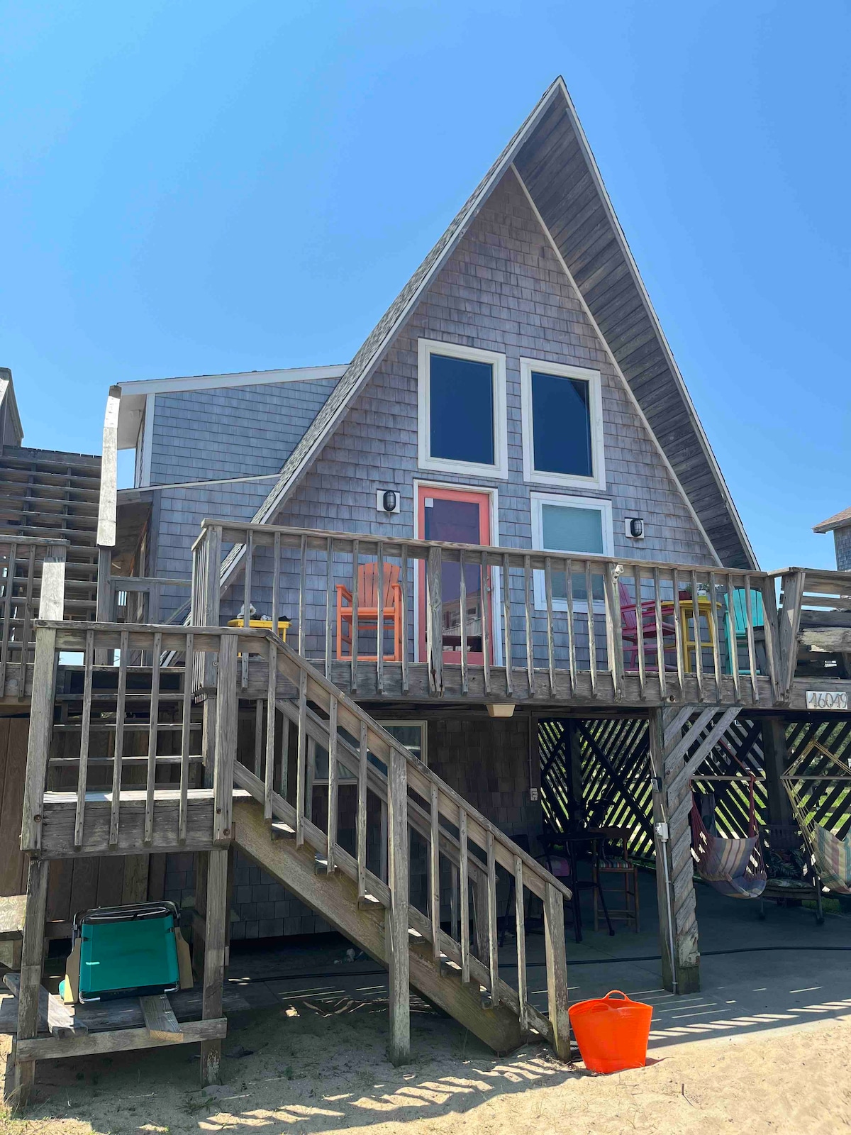The exterior of a cozy A-frame beach cottage is displayed, featuring a wooden balcony and steps leading to the entrance. Large windows offer glimpses of the interior. Brightly colored chairs are arranged on the deck, inviting relaxation. Sand is visible at the base of the structure.