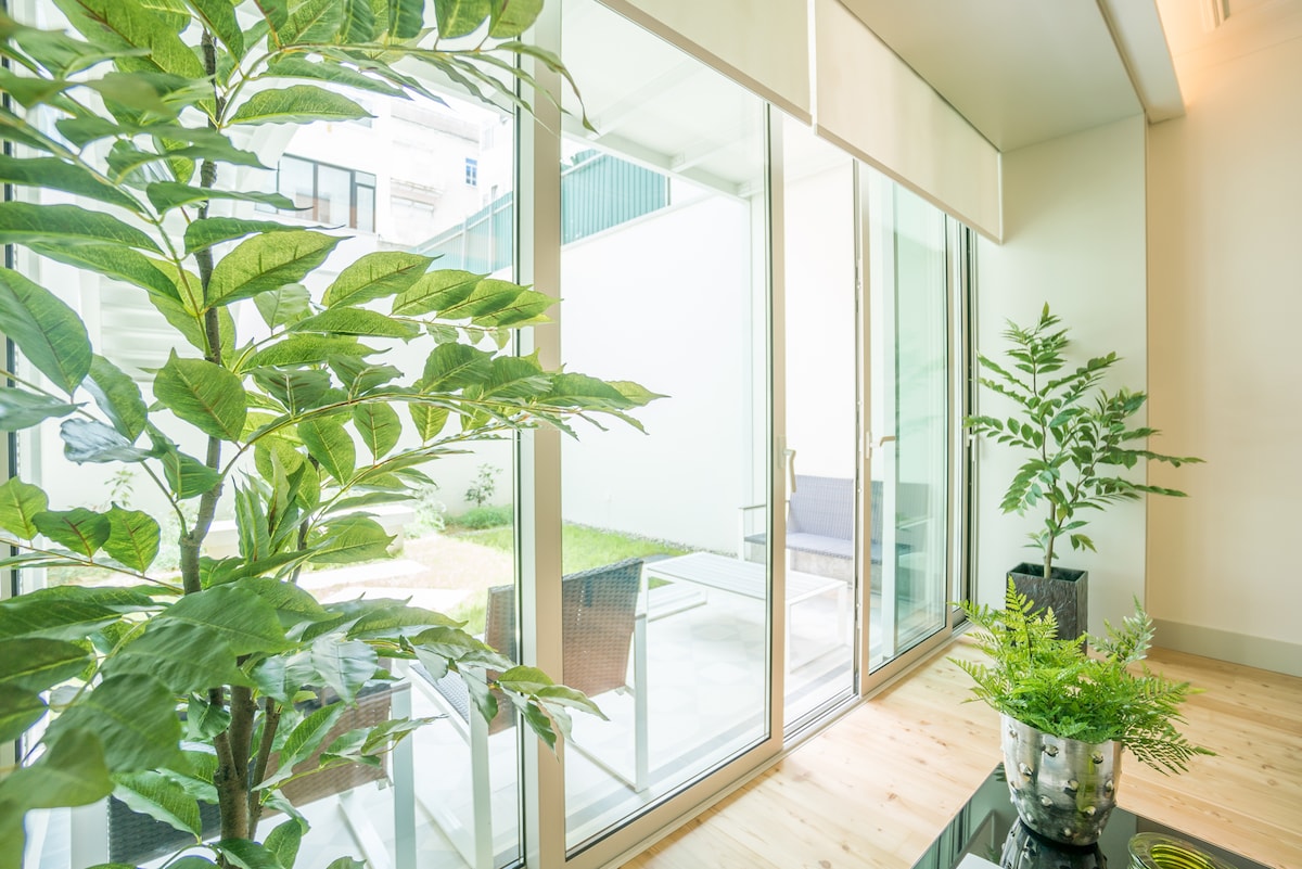 A bright and airy space is featured, with large glass doors allowing natural light to illuminate the area. Decorative plants are positioned nearby, adding a touch of greenery. A private garden can be glimpsed beyond the doors, enhancing the connection to the outdoor space.