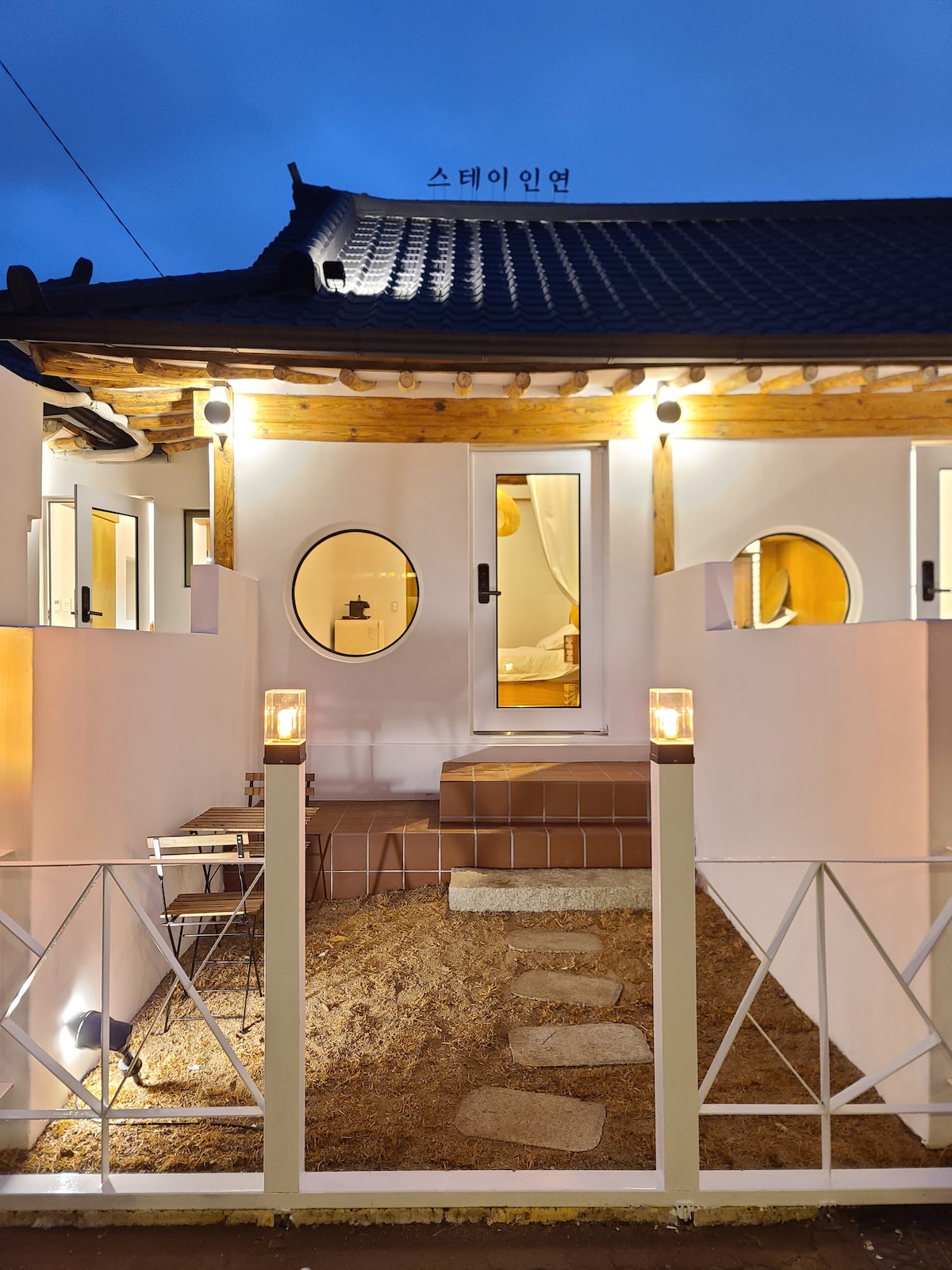 The exterior of the hanok features a welcoming entrance with steps leading to a wooden door. Circular windows and warm lighting highlight the modern design elements. A small seating area is visible, surrounded by a grassy courtyard and a decorative fence.