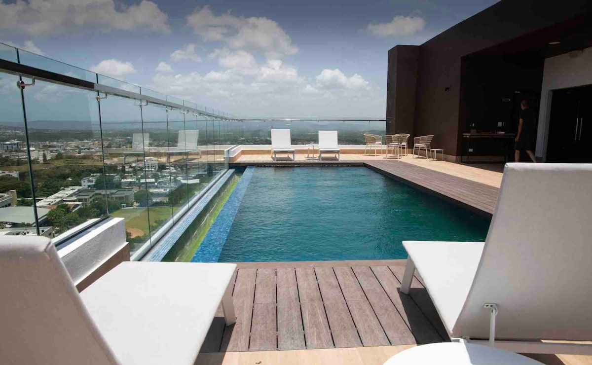 A rooftop pool is visible, featuring clear water and surrounded by a wooden deck. Two white lounge chairs are positioned nearby, offering a view of the city skyline and hills in the background. Glass railings provide an unobstructed sight of the surroundings.