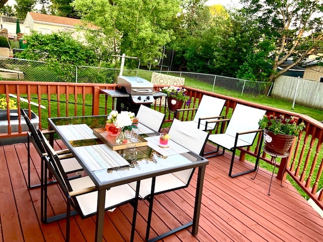An outdoor deck features a glass table surrounded by four white chairs, creating a suitable dining space. Potted plants add greenery, while a grill is positioned nearby for cooking. The backyard is partially fenced, providing a sense of privacy and a view of the lush lawn.
