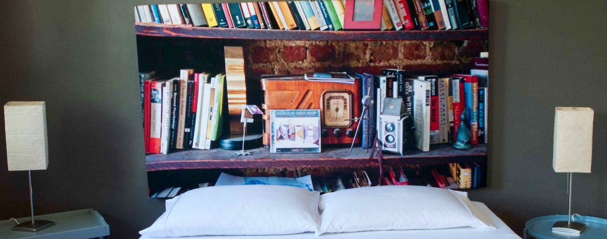 A large headboard featuring an image of a bookshelf displaying various books and vintage items, including an old radio and a film camera. Two modern bedside lamps are positioned on either side of a spacious bed adorned with white linens.