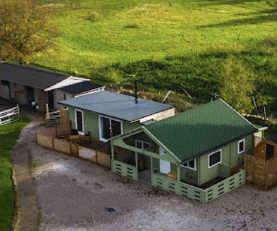 An aerial view of Fearless Fox Lodge reveals a charming green structure with a sloped roof, surrounded by lush greenery. Adjacent buildings are visible, along with a gravel path leading to the lodge, showcasing the serene, rural setting.