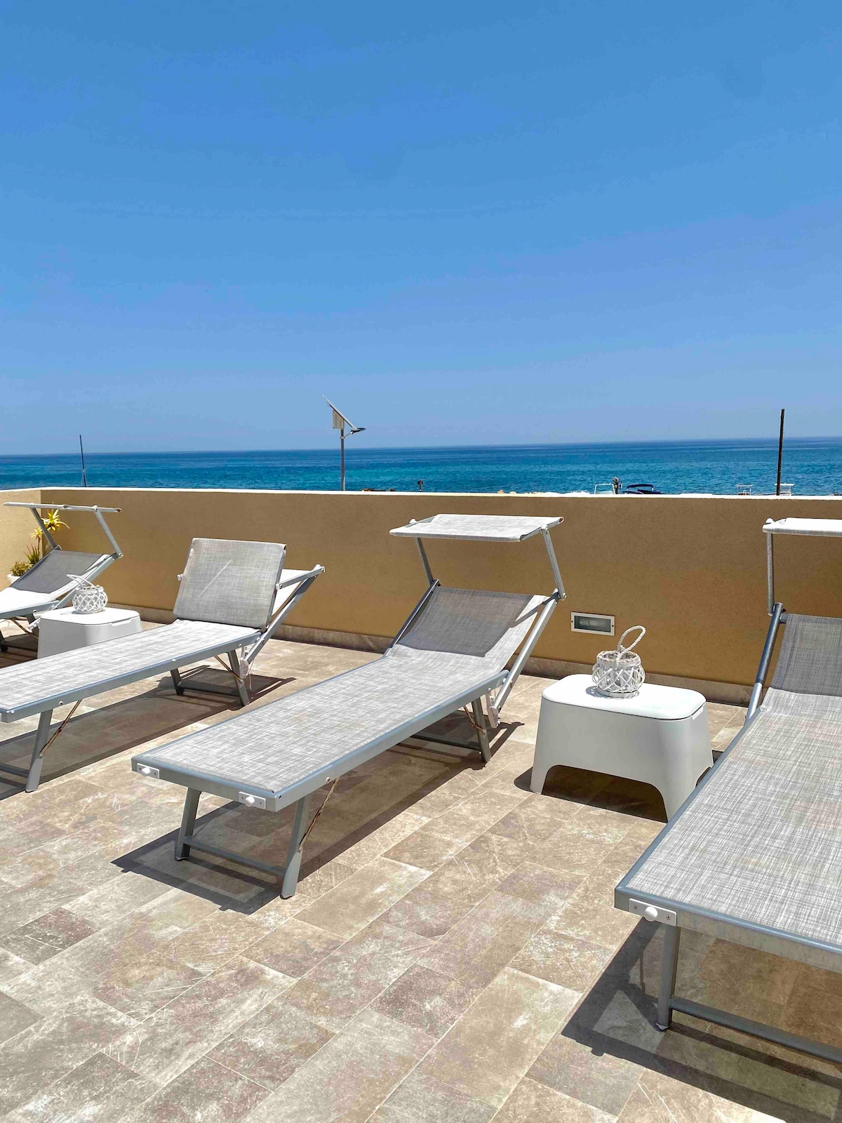 A spacious terrace is equipped with multiple sun loungers, facing a beautiful ocean view. A small side table is positioned between loungers, providing a convenient resting spot. The light-colored stone flooring complements the serene atmosphere of the outdoor space.