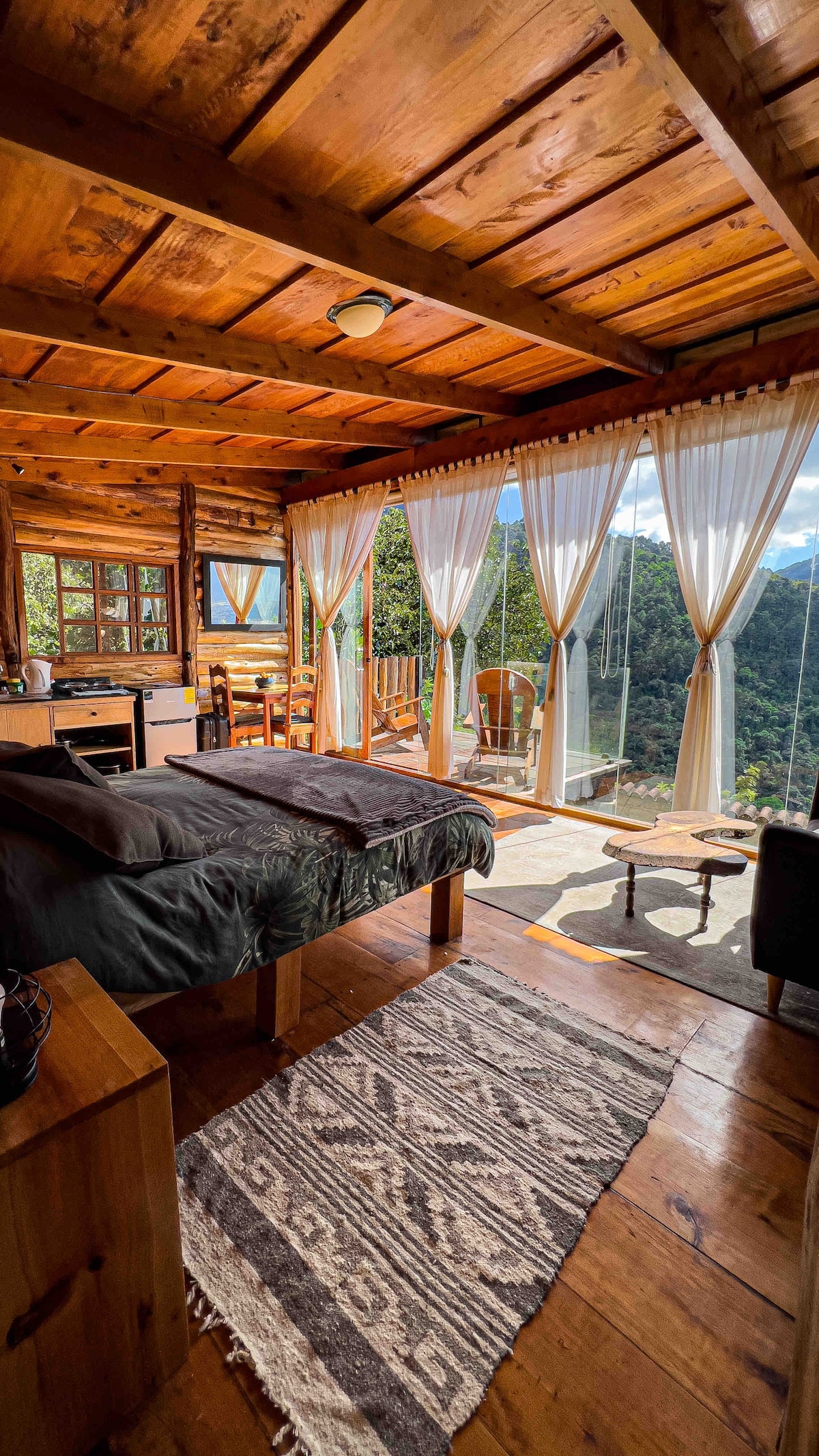 A rustic cabin interior is displayed with wooden beams and floors, accented by a cozy sleeping area featuring a dark bedding set. Natural light floods the space through full glass walls, revealing mountain views. A kitchenette and a seating area with a loveseat provide functional comfort.