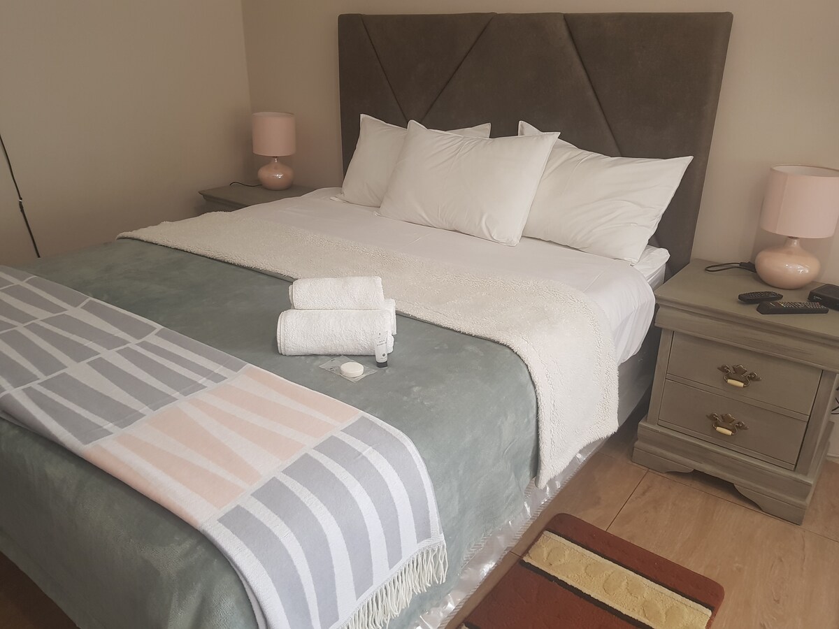 A spacious queen-sized bed is dressed with crisp white pillows and a soft, textured throw. Two bedside tables with lamps are positioned on either side. Towels are neatly arranged on the bed, complementing the serene color palette.
