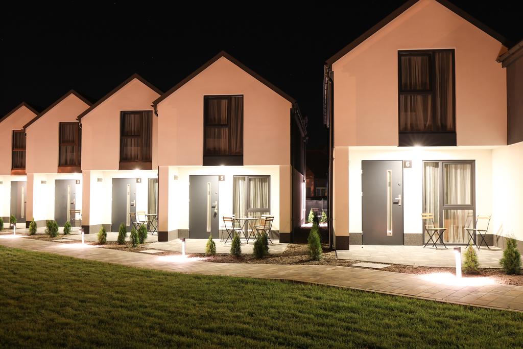 A row of modern holiday homes is illuminated at night, showcasing their clean lines and minimalist design. Each house features large windows and a front door, accompanied by small outdoor seating areas with chairs and tables. Lush landscaping accents the pathway leading to the homes.