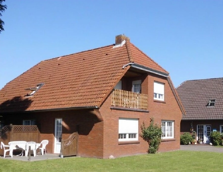 A two-story brick house is depicted with a sloped red-tiled roof. The facade includes large windows and a wooden balcony. Multiple white chairs are placed on the grass outside, surrounded by minimal landscaping and clear blue skies.