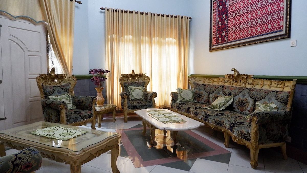 The living room features ornate furniture with intricate gold accents. A large, rectangular coffee table sits at the center, surrounded by an elaborately designed sofa set and armchairs. Natural light filters through sheer curtains, creating a warm ambiance in the space.