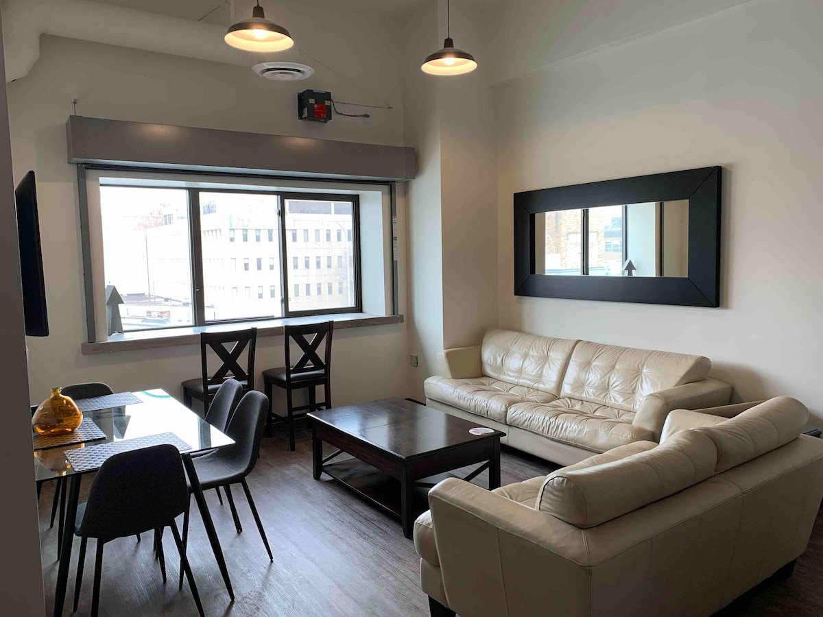 A modern living area is furnished with a white leather sofa and a wooden coffee table. A dining table with chairs is situated nearby. Large windows allow natural light to fill the space, and a wall-mounted mirror adds depth to the room.