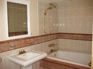 A bathroom features beige and pink tiled walls with a bathtub and a shower. A white sink is positioned beside the bathtub, and a rectangular mirror is mounted above it. Gold fixtures add a touch of elegance to the space.