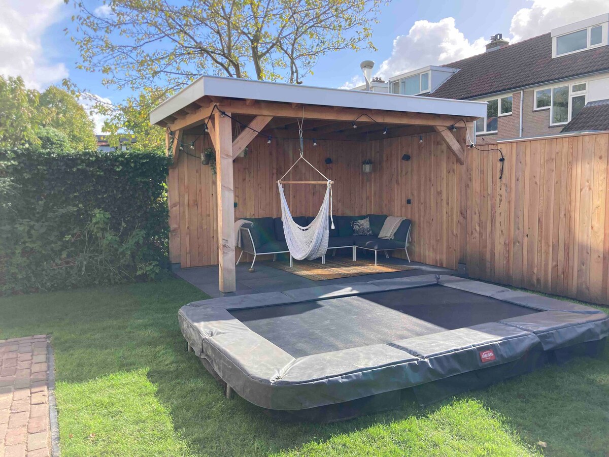 An outdoor area features a large trampoline surrounded by well-maintained grass. A wooden gazebo provides seating with a cozy hammock. The space is enclosed by a natural fence of greenery, creating a private and inviting atmosphere.