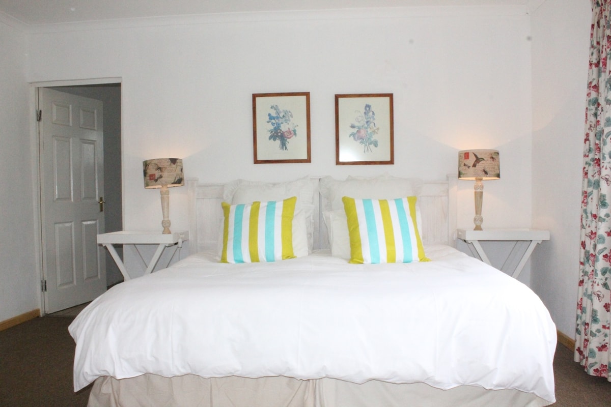 A spacious king bedroom features vibrant pillows in yellow and blue stripes on a large bed, complemented by floral artwork on the walls. Two bedside tables with lamps provide subtle lighting, while natural light enters through a partially open door leading to an adjoining room.