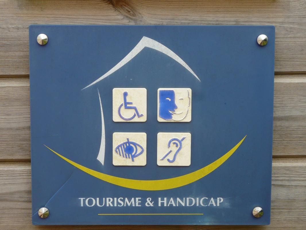 A blue sign is attached to a wooden surface, displaying symbols that denote accessibility services. The icons include representations for wheelchair access, auditory assistance, visual assistance, and mobility support, highlighting the commitment to inclusivity for all guests.