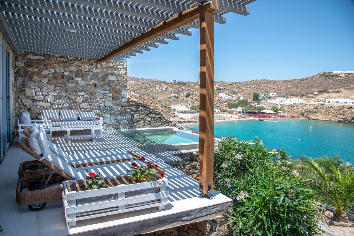 A private balcony featuring sun loungers is highlighted, overlooking a serene cove with blue waters. A swimming pool is visible adjacent to the loungers, and vibrant greenery surrounds the area. The sunlit space, framed by a wooden pergola, offers a tranquil spot for relaxation.