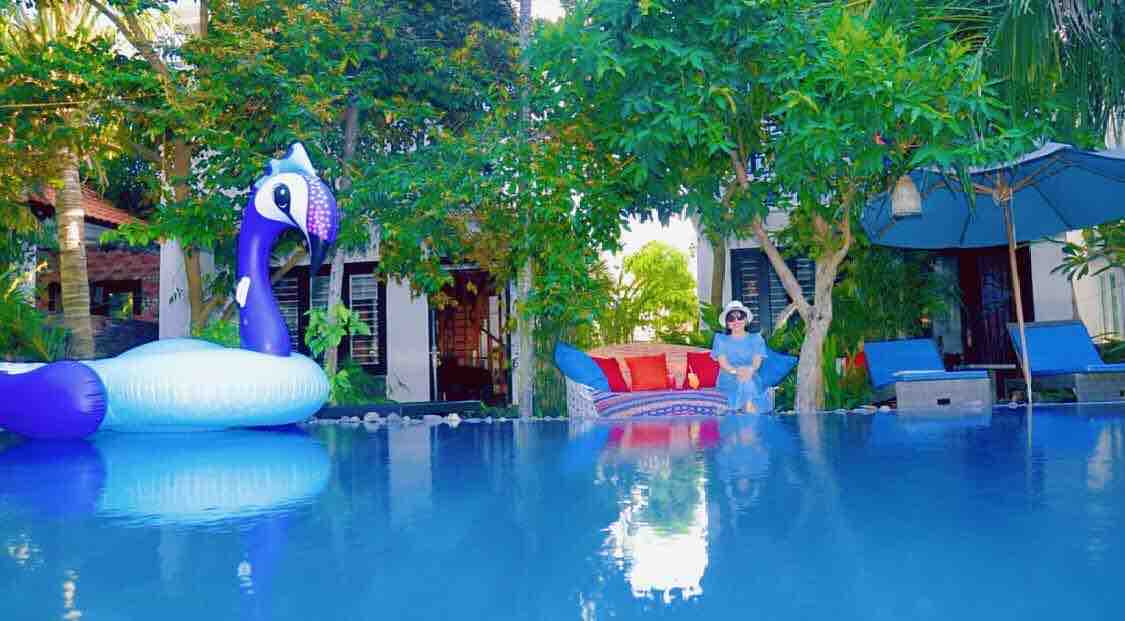 A private pool is framed by lush greenery and tropical plants. A colorful inflatable swan floats on the water, while comfortable lounge chairs and an umbrella provide shaded seating. A cozy seating area is visible in the background, enhancing the tranquil atmosphere of the outdoor space.