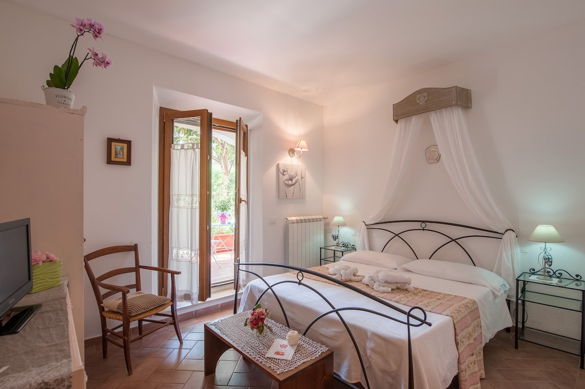A light-filled room features a spacious bed dressed in white linens, complemented by decorative pillows. Large doors open to a private terrace, with a small table and chairs visible outside. A chair and minimal decorations add a warm touch, while soft lamps provide gentle illumination.