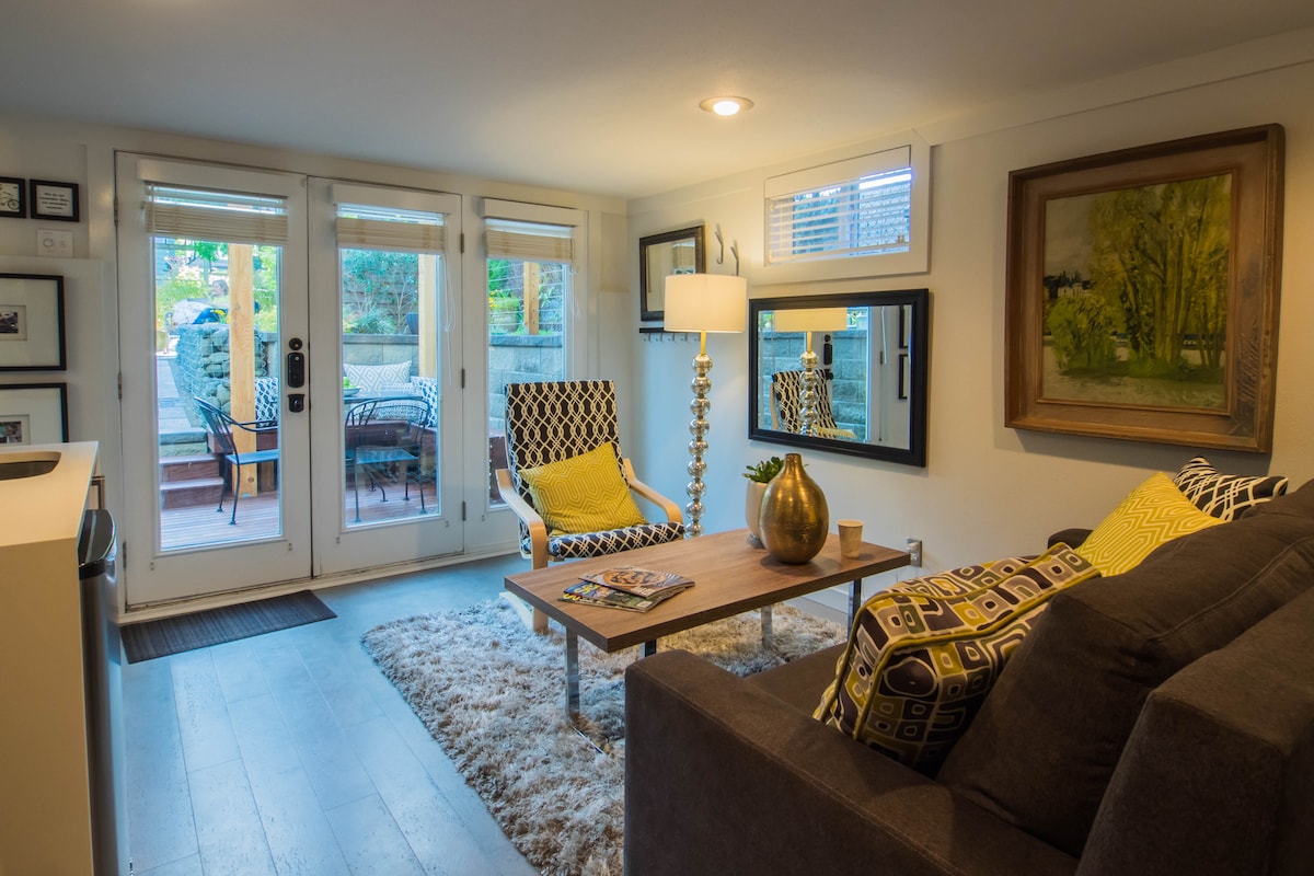 A cozy living area features modern furnishings, including a dark sofa and vibrant armchairs. A coffee table displays a magazine on a soft rug. Large glass doors provide access to an outdoor space, while natural light complements the artwork on the walls.