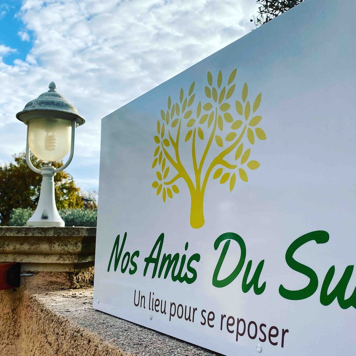 A sign displaying 'Nos Amis Du Sud' is mounted on a wall, accompanied by a decorative lamp nearby. The background reveals lush greenery and a cloudy sky, providing a calming and serene environment.
