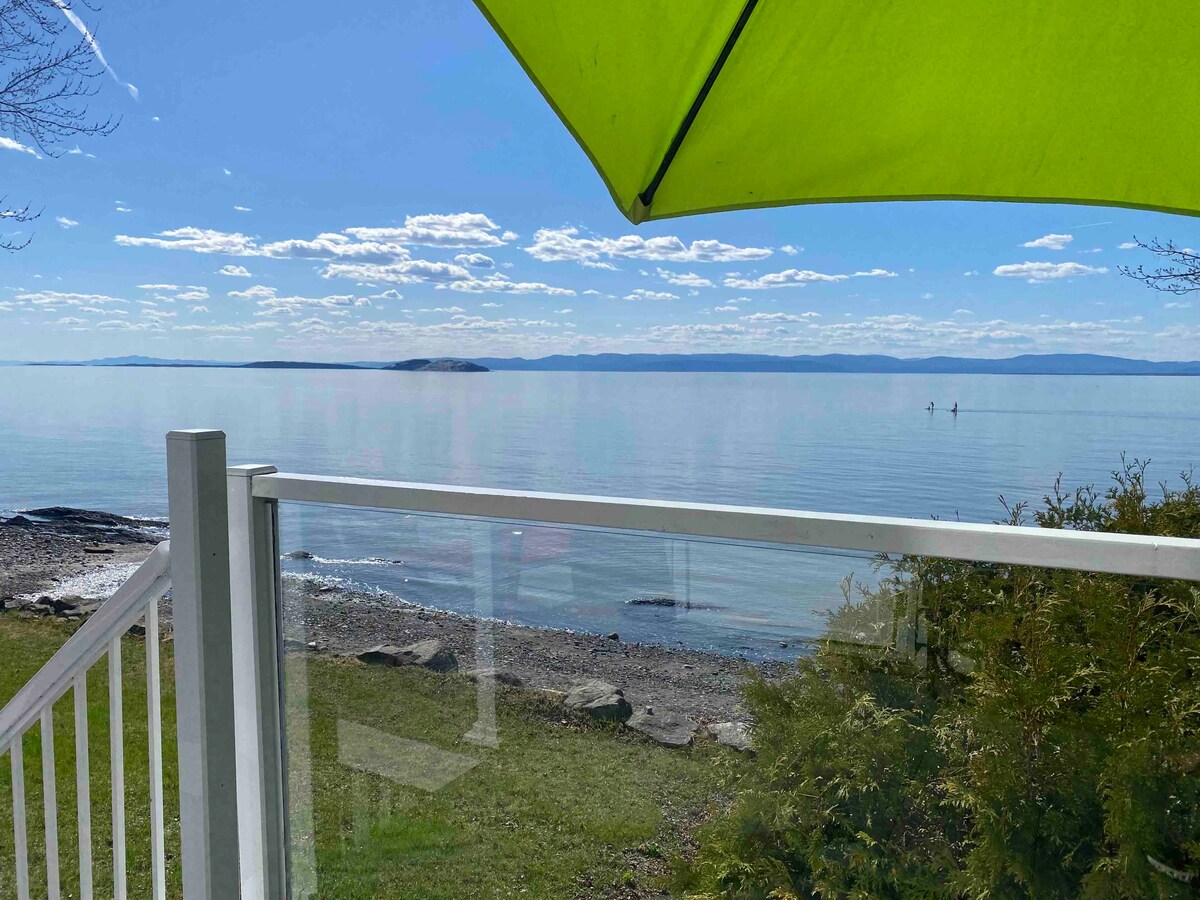 A view of calm waters extends from a spacious terrace, framed by a bright green umbrella. The shoreline is visible, with gentle waves lapping against the rocky beach. In the distance, the horizon meets the sky, showcasing a serene atmosphere.