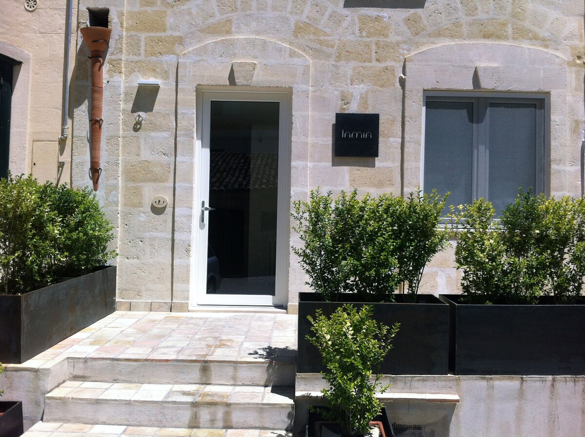 The entrance features a white door framed by stone walls, leading into the structure. Lush greenery is positioned in rectangular planters on either side, adding a natural touch to the entrance. A subtle wall plaque displays the name 'Lamia'.