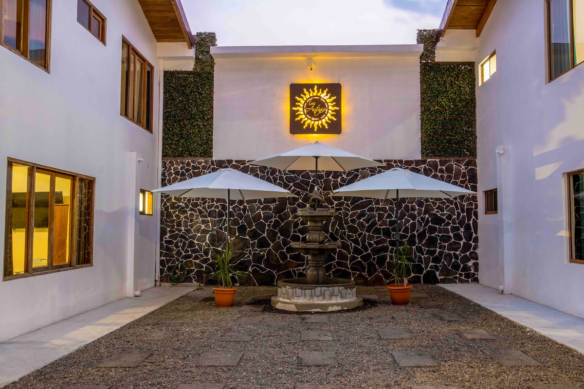 A courtyard is framed by two adjoining buildings, featuring a central stone fountain surrounded by potted plants. Two large umbrellas provide shade over a stone pathway, while walls are adorned with greenery and illuminated by a sun emblem.