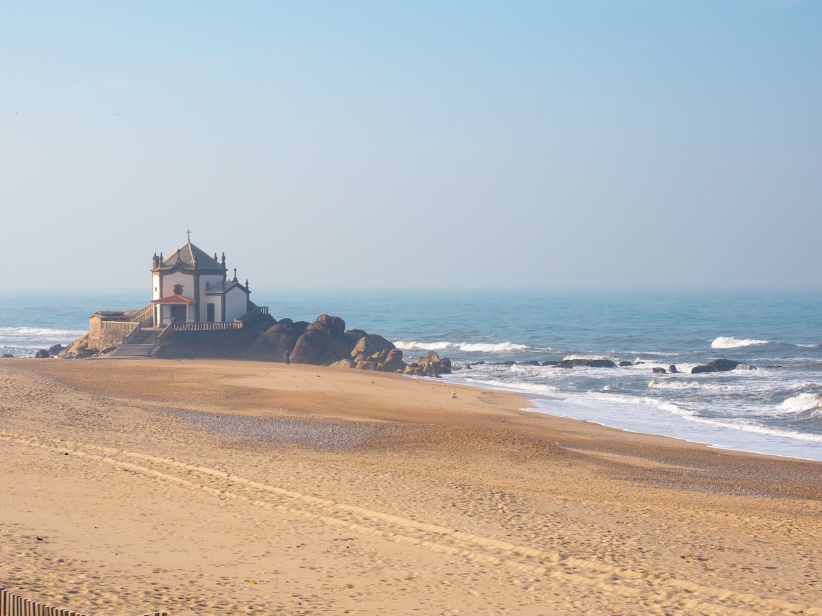 A coastal view features a small chapel perched atop a rocky outcrop, overlooking the sandy beach and gentle ocean waves. In the background, the horizon meets the sky, creating a serene atmosphere. The beach appears spacious, with footprints in the sand leading toward the water.