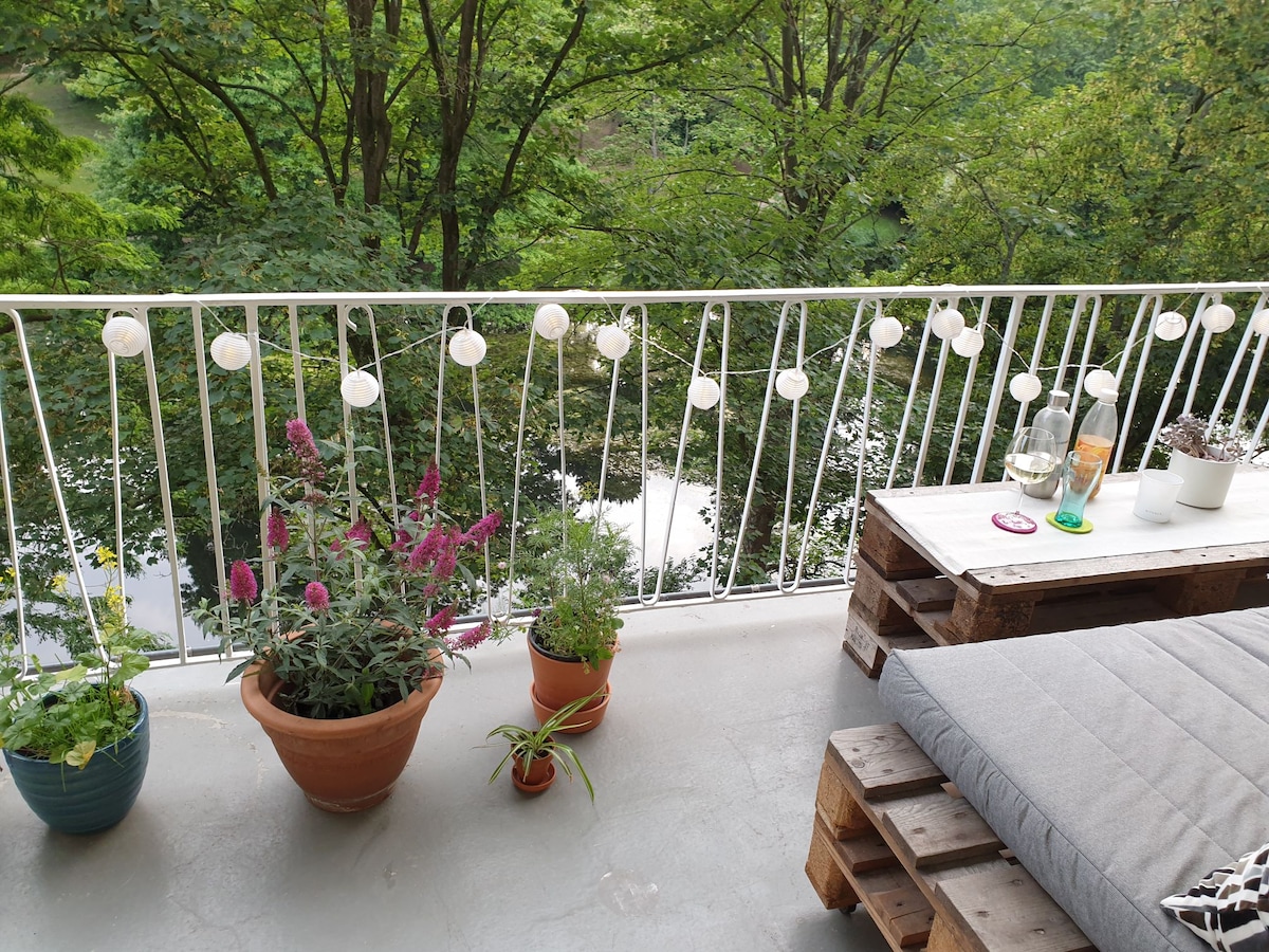 A serene balcony area features potted plants and comfortable seating arranged with wooden pallets. Soft lights are strung along the railing, providing a cozy ambiance. Green trees in the background enhance the relaxed setting, creating a tranquil outdoor space.