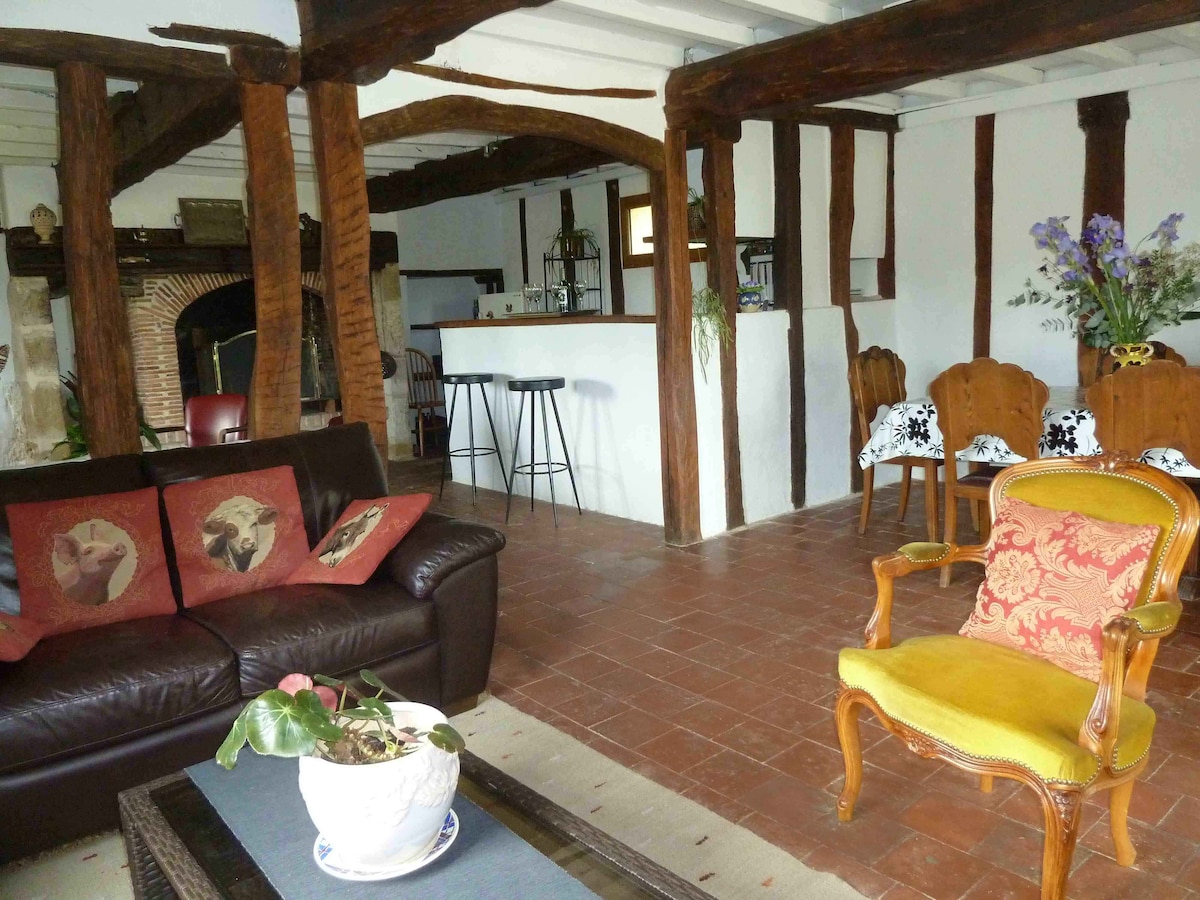 A spacious living area is highlighted by rustic wooden beams and a warm color palette. A dark leather sofa, adorned with decorative pillows, complements a vintage yellow armchair. A bar area is visible in the background, along with wooden dining chairs arranged around a table.