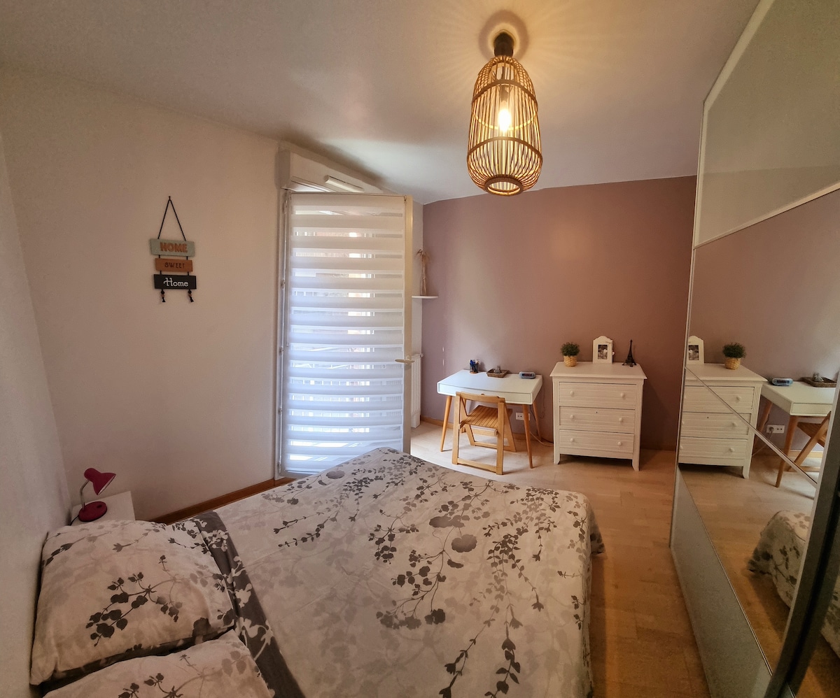 A cozy bedroom features a spacious bed with soft, floral bedding. Natural light streams through a partially closed window, and a desk with a chair is positioned nearby. A mirror and storage unit complete the minimalist decor, contributing to a calm atmosphere.