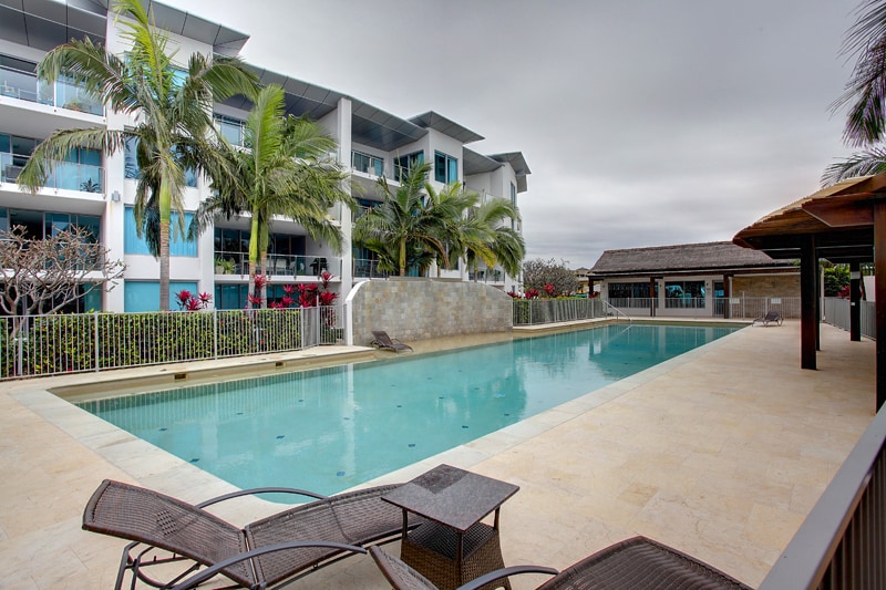 The image showcases a tranquil resort-style pool, surrounded by palm trees and modern apartment buildings. A poolside area features loungers alongside the clear water, and a shaded barbecue area is visible in the background, providing a relaxing atmosphere for guests.