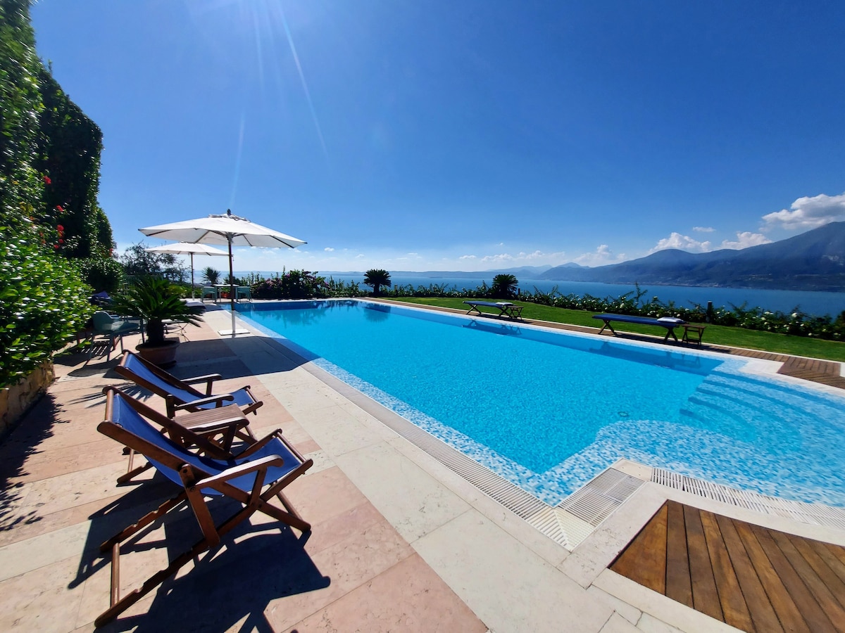 An outdoor swimming pool is surrounded by a well-maintained garden, with sun loungers arranged along its edge. An umbrella is positioned to provide shade, while a breathtaking view of the lake and mountains can be seen in the distance under a clear blue sky.