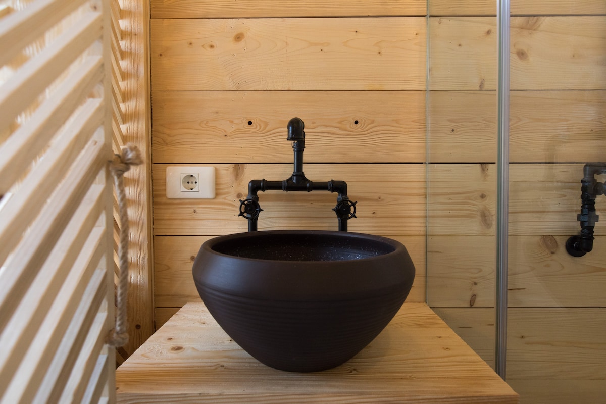 A modern bathroom features a unique, dark ceramic sink sitting on a natural wood surface. The sink is complemented by exposed black plumbing, creating an industrial contrast against the light wooden walls.